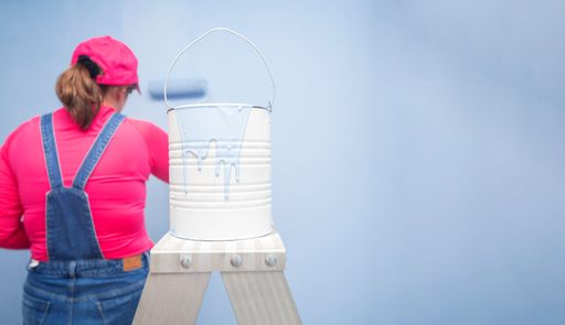 white paint can with blue paint dripping on a metal ladder, with background of a woman dressed in overalls painting with a roller a blue wall