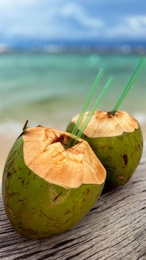 Two freshly-picked green coconuts atop a wooden table, against the sea