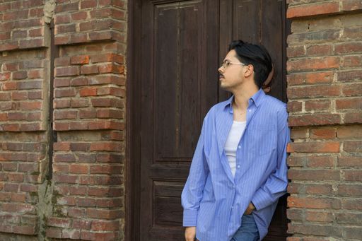 A male wearing a blue shirt, leaning against a brick wall and a wooden door