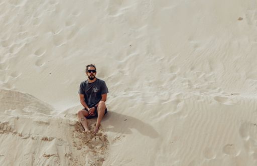 A Hispanic young surfer man with sunglasses sitting on sand dunes