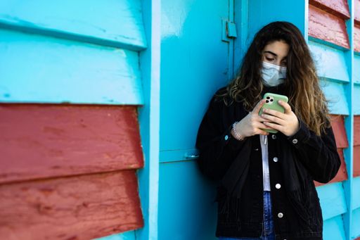 A Caucasian woman with protective mask browsing her phone while standing against a blue metal door