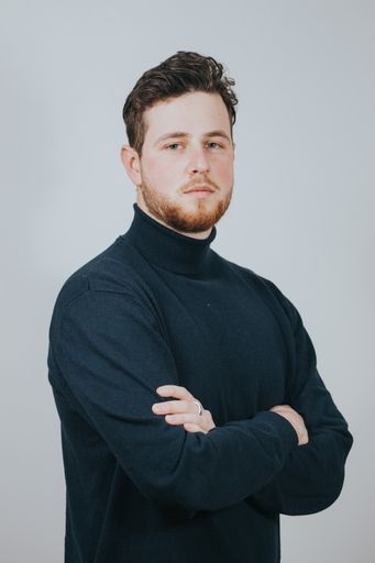 A vertical shot of a young Caucasian man wearing a turtleneck, with his arms crossed in front of a white background