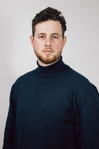 A vertical shot of a young Caucasian man wearing a turtleneck, looking serious in front of a white background