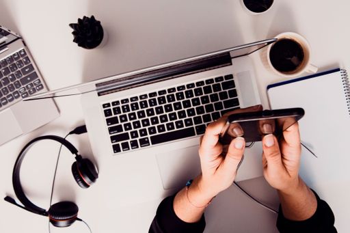 A man using a smartphone on a table next to an open laptop with headphones