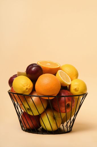 A black metal basket filled with a range of fresh, juicy fruits against a peach-colored background