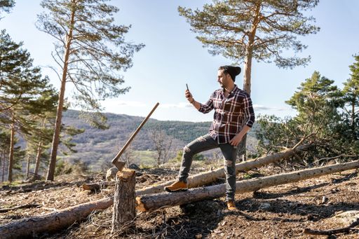 A closeup of a lumberman taking a photo with a smartphone next to his ax stuck on the stump