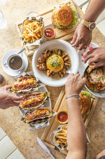 A top view of the hands of people grabbing food from the table served with hamburger, tacos, fries and salads