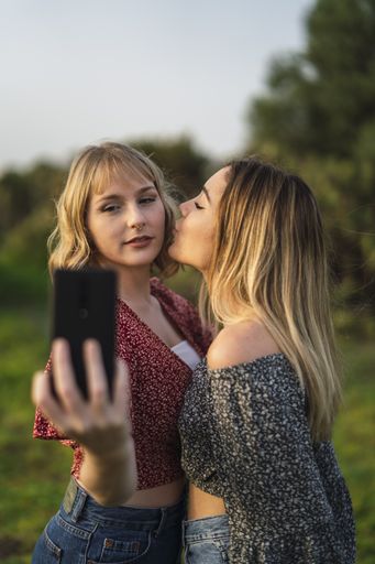 A vertical shot of two beautiful Spanish Caucasian friends taking a selfie in the park, one of them kissing the other