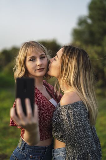 A vertical shot of two beautiful Spanish Caucasian friends taking a selfie in the park, one of them kissing the other