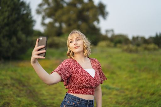 A shallow focus of a young blonde woman wearing a floral crop top and jeans and taking a selfie in a park