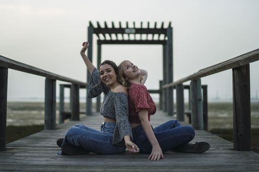A shallow focus of two cheerful female friends sitting on a wooden deck in a field in Spai