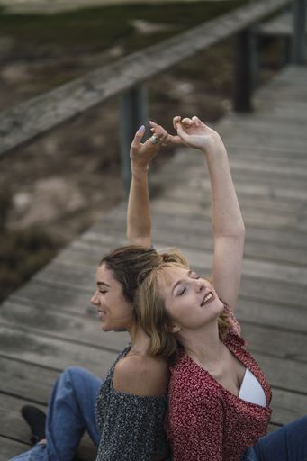 A vertical shot of two beautiful Spanish Caucasian girls sitting back to back on a wooden pier