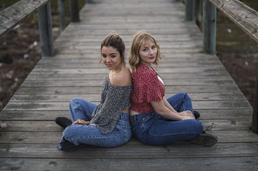 A shallow focus shot of two beautiful Spanish Caucasian girls sitting back to back on a wooden pier