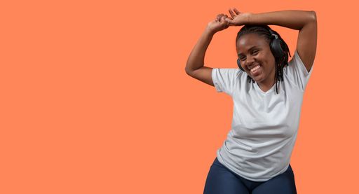 young African woman dancing and listening to music with headphones, arms up, dressed in white t-shirt and blue pants against orange colored background