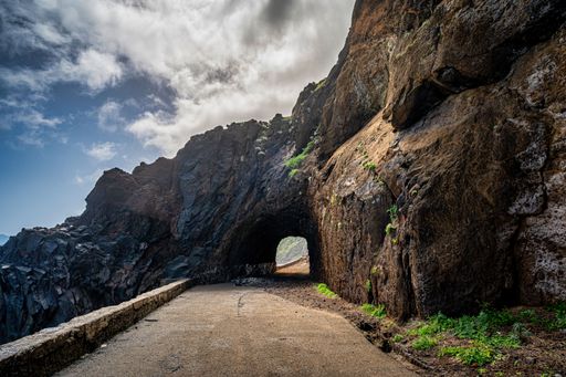 A scenic view of a winding mountain road passing through an underground tunnel lit by sunlight, with a blue sky in the background