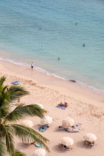 An aerial view of a sandy beach with crystal clear water. The people on the beach and in the sea are tiny compared to the vastness of the scenery.