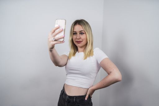 A young Caucasian female from Spain taking a selfie with a phone against a white wall