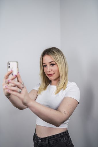 A young Caucasian female from Spain taking a selfie with a phone against a white wall