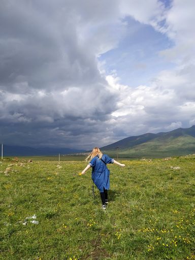 A vertical shot of a blonde-haired female dancing in a large beautiful green field with breathtaking clouds in the sky