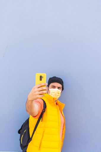 Spanish man with face mask taking a selfie with the yellow mobile phone on a blue background