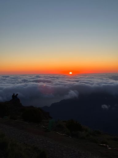 A group of people stands on high cliff overlooking the horizon with the sun setting in the background