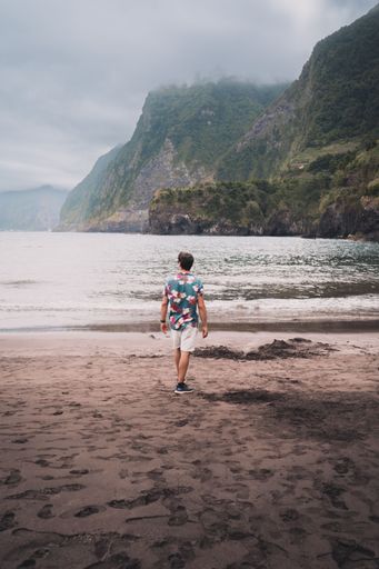A man in casual shorts is strolling along a sandy beach, with a beautiful view of green mountains.