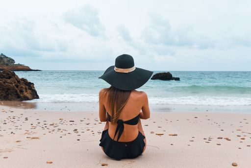 A young woman wearing a wide-brimmed hat sits on a sandy beach overlooking the calm blue ocean.