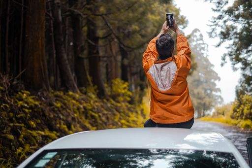 A man in an orange jacket sitting on the roof of a car and taking pictures with his phone.