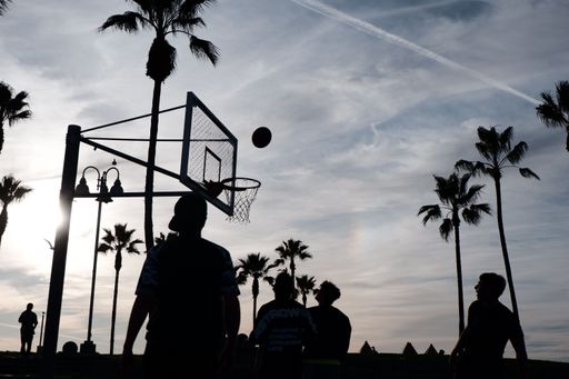 The silhouettes of a group of people enjoying a game of basketball at an outdoor court surrounded by palm trees on a sunny day