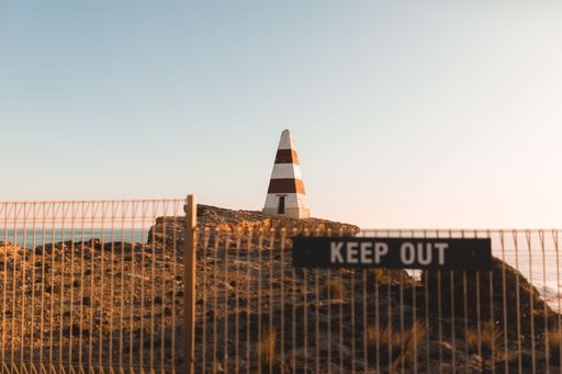 A fence with a 'Keep Out' sign situated in front of a lighthouse in the distance, against an empty sky