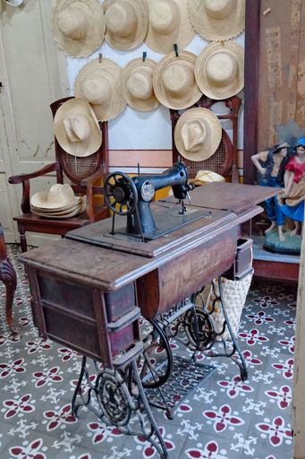 A vertical shot of a tailor's studio with a sewing machine and hats hanging on the wall