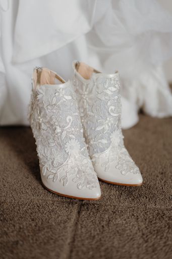 A pair of pristine white wedding shoes on a brown carpet floor with a wedding dress in the background