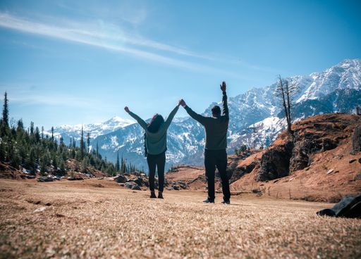A back view of travelers with raised hands facing a scenic mountainscape, Kothi Meadows, Manali, India