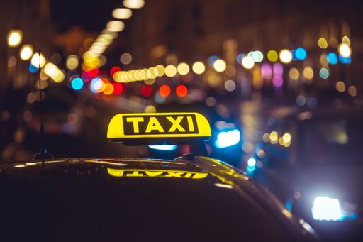 Illuminated taxi sign on top of a car. City lights with neon color in the background. Rainy urban night scene.