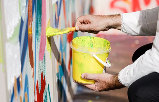A closeup of a hand of a person holding yellow paint and brush and painting a colorful wall