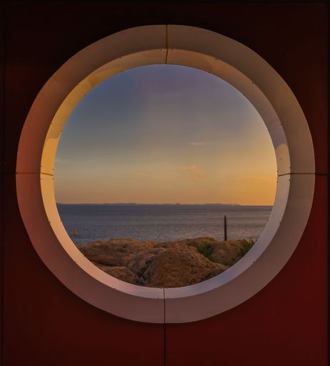 A round window from the inside of a room with the sea at sunset in the background
