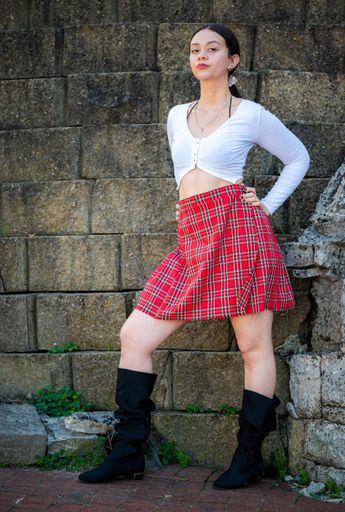 A young woman posing against a stone building, wearing a red checkered skirt