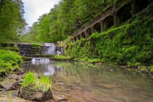 A picturesque waterfall cascading down a rocky cliff face surrounded by lush greenery, towering trees and a bridge in a peaceful forest setting