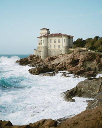 An impressive white, old castle in Italy atop a cliffside, with waves crashing on the shore