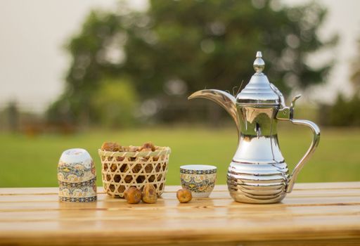 A silver teapot on a wooden table with cups and dried apple on a grassy lawn in a sunny outdoor setting