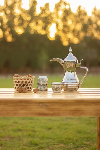 A silver teapot on a wooden table with cups and dried apple on a grassy lawn in a sunny outdoor setting