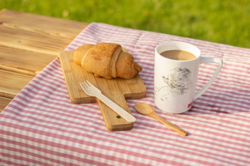 A wooden table with a white porcelain coffee cup, a croissant, spoon, fork on a sunny outdoor setting