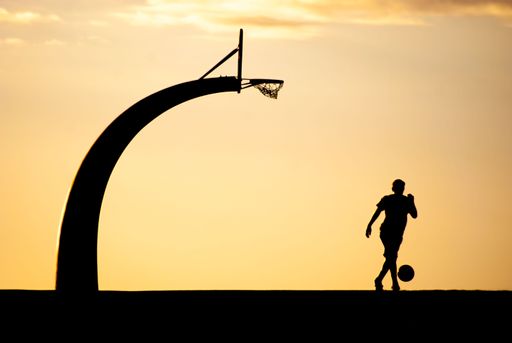 A silhouette of a man with a basketball hoop and ball against a backdrop of an orange sky at sunset