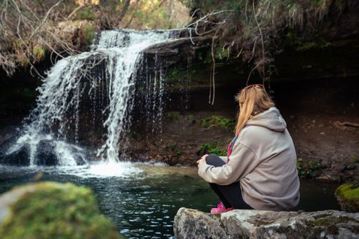 A person seated on rocks gazes at water cascading from a waterfall