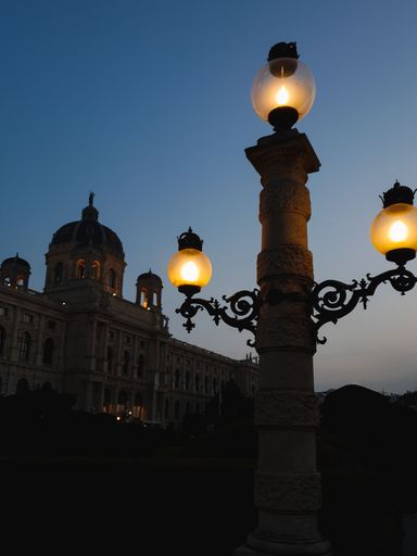 An isolated, vintage street lamp stands alone in front of the Natural History Museum of Vienna, Austria, illuminated by the setting sun