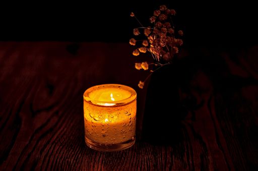 An illuminated candle atop a wooden table with dried flowers over it in a dark room