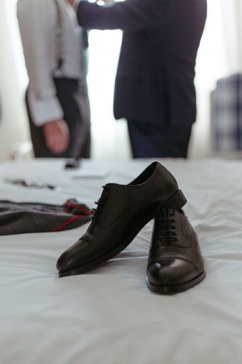 Black wedding shoes on a bed of white linen, with men in a classic tuxedos in the background