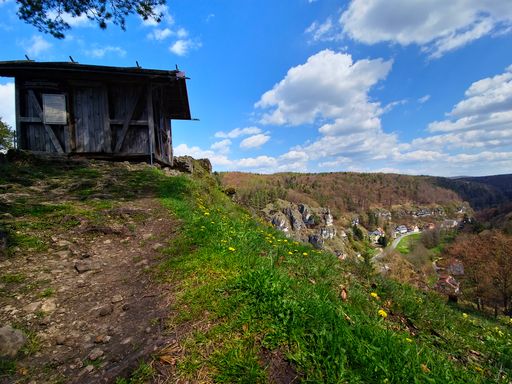 A rustic wooden cabin sits atop a grassy hill, surrounded by lush trees and a breathtaking valley in the horizon