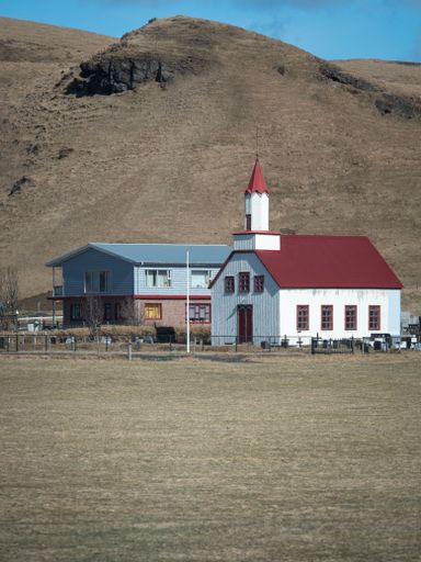 A church stands alone in a vast, grassy field, framed by majestic mountain peaks in the background