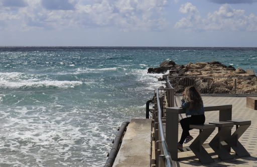 A Caucasian girl sits at a wooden table on a pier watching the foamy sea in a bright windy day